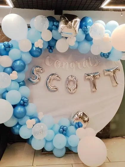 A custom "Congrats" backdrop designed to celebrate a personal or professional achievement. The circular backdrop is framed with a blue and white balloon garland and features silver foil letter balloons to spell out a name or message.