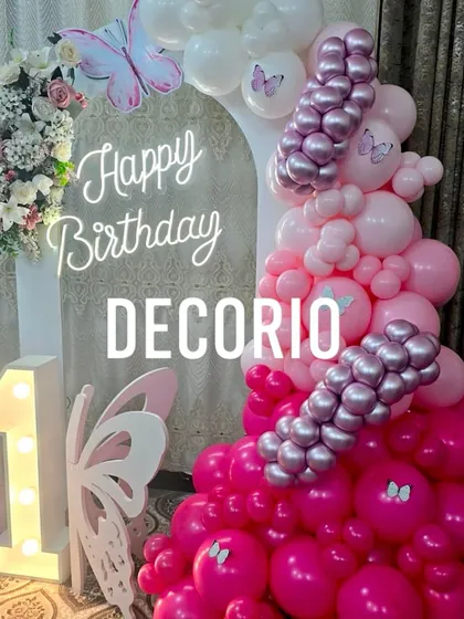 A first birthday celebration with a vibrant butterfly theme. This setup includes a '1' marquee number, a circular backdrop with a neon sign, and a stunning balloon arrangement in shades of pink and purple with butterfly details.