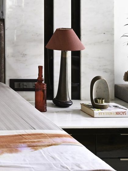 This bedside vignette combines function and style. A sleek table lamp provides reading light, while decorative objects and a potted plant add color and life against the backdrop of a white marble feature wall with black vertical grooves.