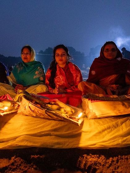 A family sits by the riverbank at night, their faces illuminated by the warm glow of diyas as they wait for sunrise during Chhath Puja.