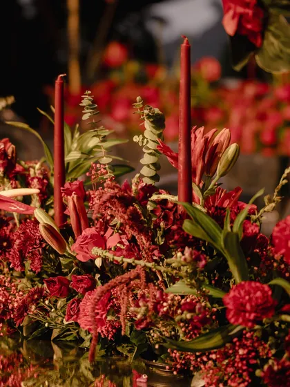 A close-up of the centerpiece for the red Sangeet. The arrangement features a mix of red lilies and other blooms, with tall red taper candles adding height and a warm glow.