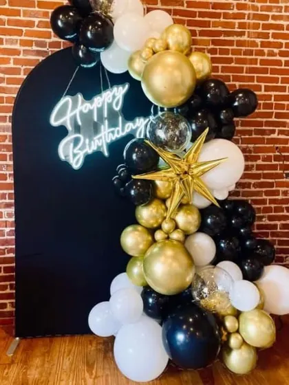 A close-up of a chic black, gold, and white balloon garland against a black backdrop, complete with a "Happy Birthday" neon sign.