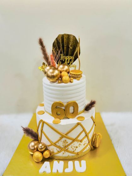 An elegant two-tiered cake for a 60th birthday. The design features clean white frosting, a modern geometric gold pattern, and is topped with dried botanicals and macarons.