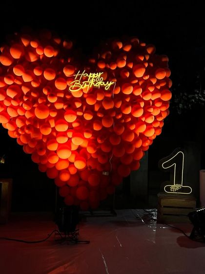 A dramatic and unique first birthday decoration featuring a giant heart made of red balloons, illuminated from within, with a neon number '1' on the side.