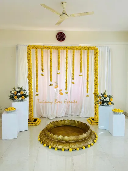 Our simple Haldi setup is perfect for any space. It includes a marigold arch, a large brass Urli for the ceremony, and two white pedestals with floral arrangements to frame the space beautifully.