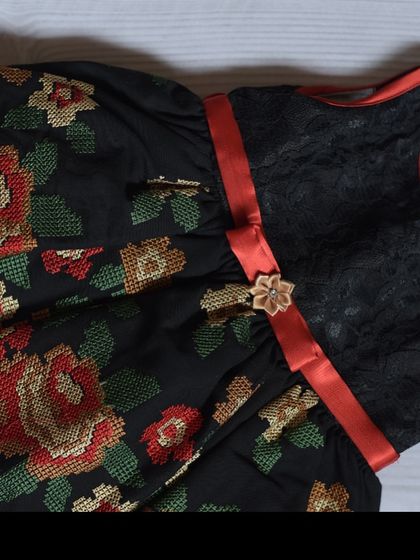 This unique dress features a black lace bodice paired with a skirt made of cross-stitch style floral embroidered fabric. A simple red ribbon ties the bold look together.