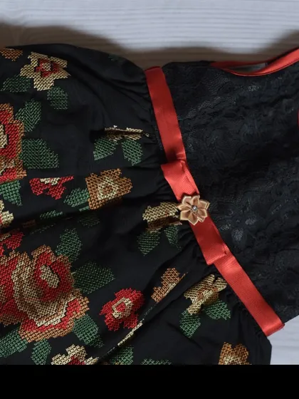 This unique dress features a black lace bodice paired with a skirt made of cross-stitch style floral embroidered fabric. A simple red ribbon ties the bold look together.