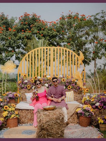 The couple posing at their rustic Haldi seating area. The backdrop of a yellow slatted arch, hay bales, and an abundance of sunflowers creates a cheerful and picturesque setting.