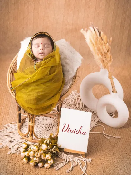 A simple and elegant newborn portrait. The baby is wrapped in a soft green cloth, resting in a cane chair with a personalized name card.