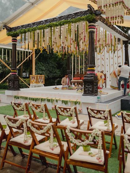 The outdoor mandap setup with traditional dark wood pillars, adorned with hanging floral strings of tuberose and lotus buds. The seating is arranged for an intimate ceremony.