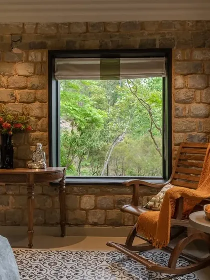 A cozy reading corner in one of the bedrooms, featuring a rocking chair and large windows that frame the lush greenery. The exposed local stone wall adds a rustic and authentic touch.