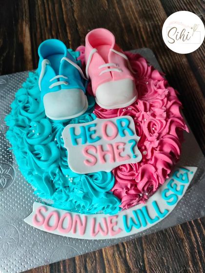 "He or She? Soon we will see." This gender reveal cake features cute fondant baby shoes in pink and blue, sitting on a bed of matching buttercream rosettes.