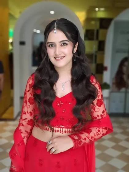 Radiant in red. This engagement look features a bright red lehenga, soft curls, a delicate maang tikka, and makeup that is fresh, young, and glowing.