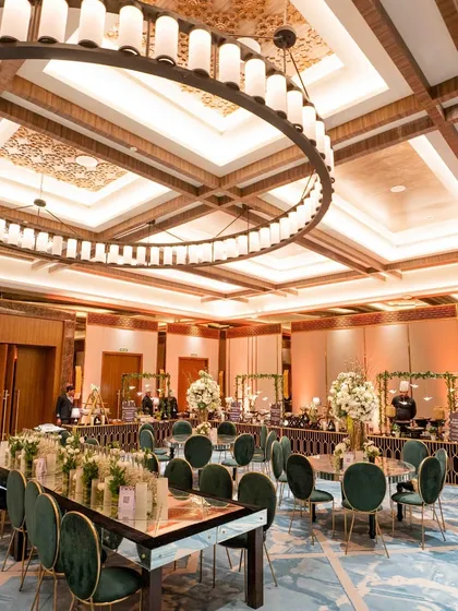 A modern and luxurious dining setup with a U-shaped table, emerald green velvet chairs, and a massive circular chandelier.