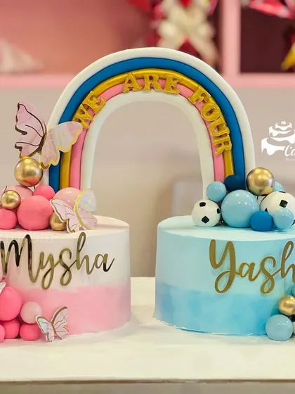 A creative cake set for twins or a baby shower. Two separate cakes, one pink for a girl and one blue for a boy, are connected by a rainbow that says 'We Are One'.