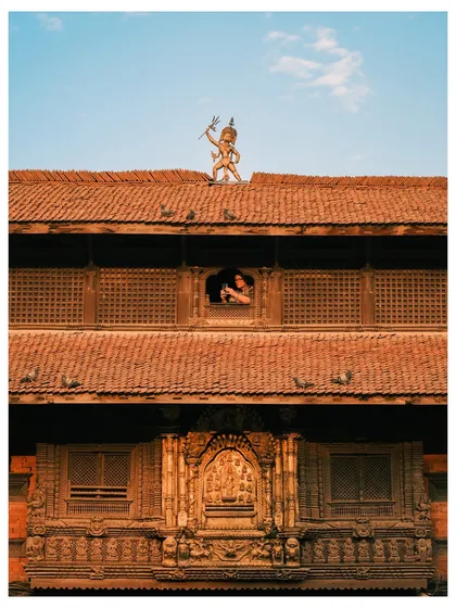 A unique view of a traditional building in Kathmandu, Nepal. The frame captures a deity statue on the roof and a person looking out from a beautifully carved wooden window.