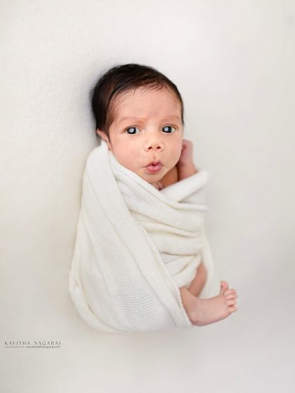 A newborn wrapped in a simple white cloth, looking curiously at the camera. The high-key lighting and minimalist approach create a clean and modern portrait.