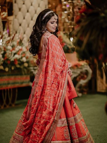A full-length portrait of the bride in her beautiful orange lehenga. The garden setting and her graceful pose create a classic and elegant bridal shot.