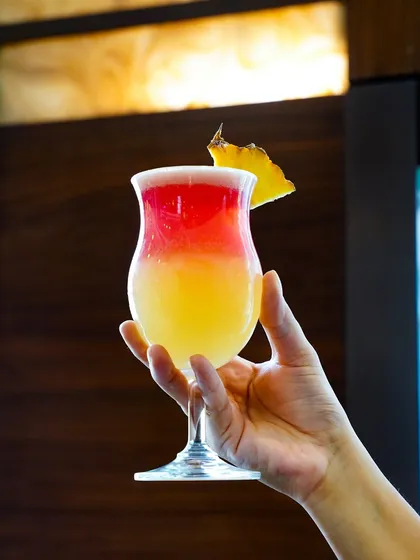 A hand holds a layered tropical cocktail with red and yellow hues against a wooden background. This image is perfect for showcasing a signature drink on a restaurant or bar menu.