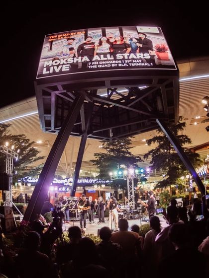 A wide shot showing the band on a large outdoor stage with a big crowd and a huge screen with "Moksha All Stars Live". This communicates scale and experience with large events.