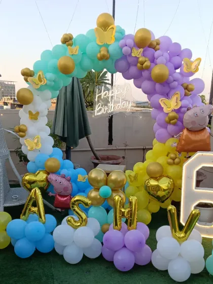 A colorful Peppa Pig themed party set up on a green terrace. I created a multi-color balloon ring arch and spelled out the name "KASHVI" with individual foil balloons at the base. The setup also includes a Peppa Pig foil balloon and a light-up number '6'.