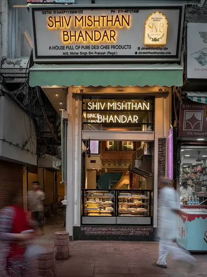 A long-exposure shot of the Shiv Mishthan Bhandar storefront, capturing the motion of the bustling street. The well-lit interior acts as a beacon, drawing customers in from the narrow lane.