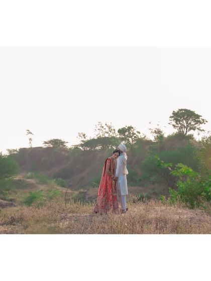 A wide shot of a couple in their wedding attire, resting against each other in a quiet, natural landscape, taking a peaceful moment together.
