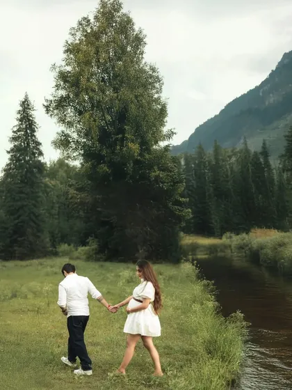 Walking together along the river. This shot captures the couple in motion, hand-in-hand, with the beautiful natural scenery of the forest and mountains creating a peaceful backdrop.