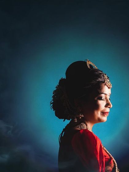 An artistic bridal portrait against a moody, blue-toned background. The light creates a halo effect around her, highlighting her traditional hairstyle and serene, closed-eyed expression.