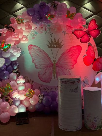 This butterfly-themed first birthday is all about enchantment. I used a circular backdrop with a large butterfly graphic, surrounded by a lush balloon garland in pink and purple, and accented with large 3D butterfly props and a lit-up number '1'.