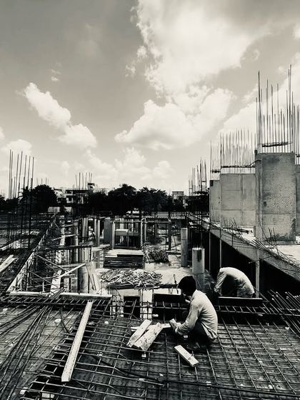 A black and white photograph from a construction site, capturing workers against a backdrop of concrete pillars and rebar. The image powerfully quotes Winston Churchill: "We shape our buildings; thereafter, they shape us."