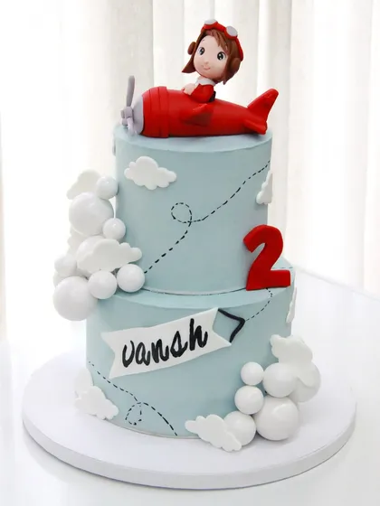 Fasten your seatbelts! This two-tiered airplane cake celebrates Vansh's 2nd birthday with a cute pilot, fluffy clouds, and a sky-blue ganache finish.