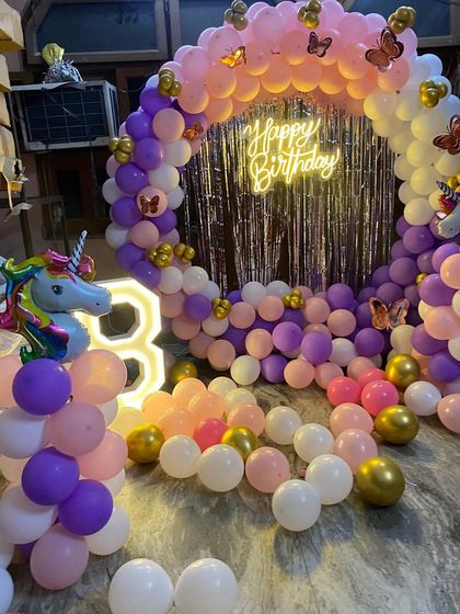 A magical unicorn-themed 3rd birthday party. The decor includes a circular balloon arch in pink, purple, and gold, unicorn foil balloons, and a light-up number '3'.
