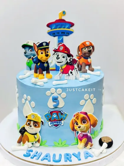 A cute Paw Patrol theme cake with 2D pup characters. The cake is decorated with edible paw prints and bones, with the iconic lookout tower on top.