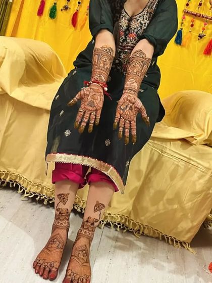 A bride sitting on a decorated seat at her mehendi ceremony, displaying her full hand and foot mehendi.