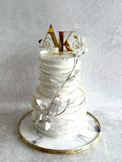 This two-tiered wedding cake features a modern, textured ruffle design. The clean white finish is accented with delicate, silver-dusted leaves and a custom gold initial topper, creating a sophisticated centerpiece.