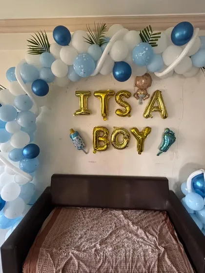 An 'It's a Boy' welcome home decoration featuring a blue and white balloon half-arch over a sofa, with gold foil letters.