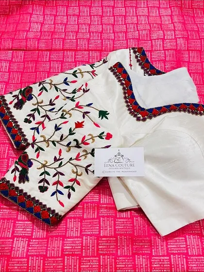 This elegant white blouse features colorful bird and floral motifs embroidered with threadwork. The neckline and sleeves are highlighted with a traditional red and blue border, perfect for pairing with a classic Kanchipuram saree.