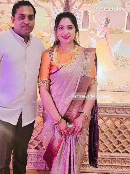 A simple and elegant coordinated look. She wears a beautiful silk saree with a custom orange blouse featuring detailed embroidery on the sleeves, while he wears a classic white shirt.