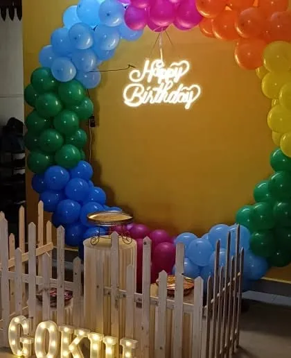 A bright rainbow-colored balloon ring with a neon sign, enclosed by a white picket fence.