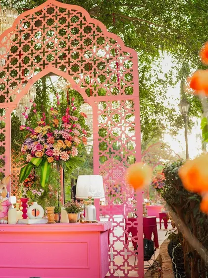 This vibrant pink bar and photo booth, framed by a beautiful jaali arch, serves as a stunning focal point. It's decorated with lush flowers and quirky details for a fun and festive vibe.