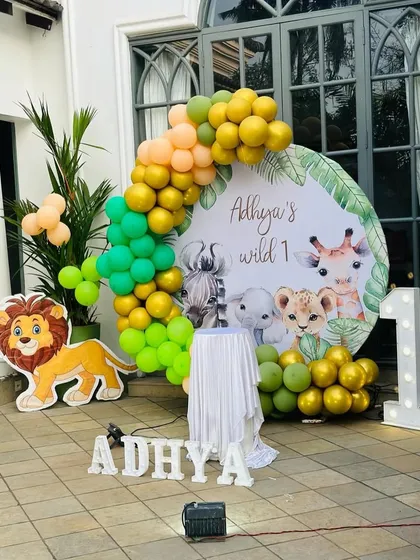 A "Wild One" jungle safari theme for a first birthday. This outdoor setup includes a circular backdrop with cute animals, a lion cutout, and a green and gold balloon arch.