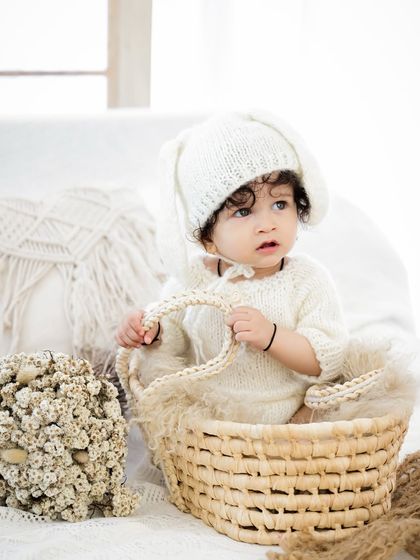A curious little bunny. This baby is wearing a cute knitted outfit with bunny ears, sitting in a basket with natural, dried flowers.
