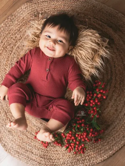 A happy baby in a maroon romper lies in a wooden crate on a jute rug. The simple, rustic props and pops of red berries create a warm and earthy portrait.