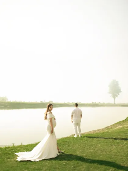 An elegant and serene portrait by the water. The mother-to-be, in a flowing white gown, looks back at the camera while her partner stands contemplatively, creating a timeless and artistic image.