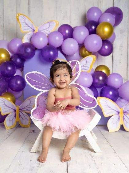 A sweet toddler dressed as a butterfly, smiling brightly for her milestone photoshoot. The purple and gold balloon arch and butterfly cutouts create a magical, whimsical setting.
