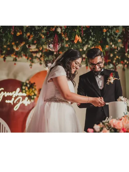 The happy couple cutting their cake at a reception with a vibrant, fall-colored theme. The backdrop features a custom neon sign with their names and a ceiling lush with hanging florals and disco balls.