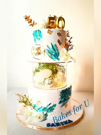 A luxurious two-tier 50th birthday cake with a clear, illuminated separator. The design features abstract teal brushstrokes, gold leaf, and delicate white flowers.