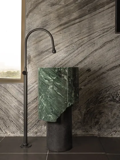 We see furniture and fixtures as sculptural elements. This freestanding pedestal sink, custom-crafted from a single piece of green marble with a raw, broken edge, stands as a piece of art against the dramatic stone wall, paired with a minimalist matte black faucet.
