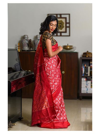 A side profile shot that highlights the beautiful drape of the red saree and the complementary brocade blouse. This image focuses on the complete ensemble in a refined, indoor lifestyle setting.